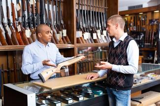 Man behind a counter displaying firearms conversing with another man.